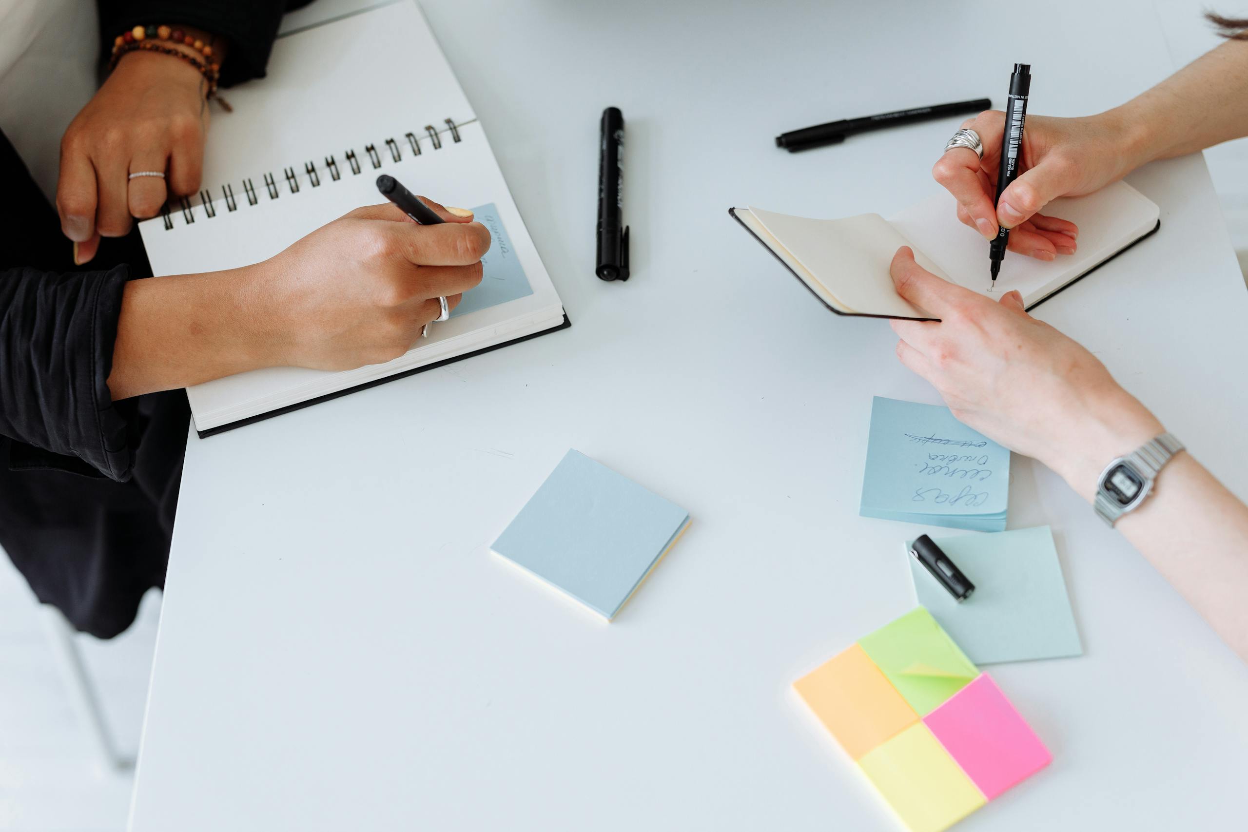 Top view of people collaboratively writing notes on a desk with notebooks and sticky notes.